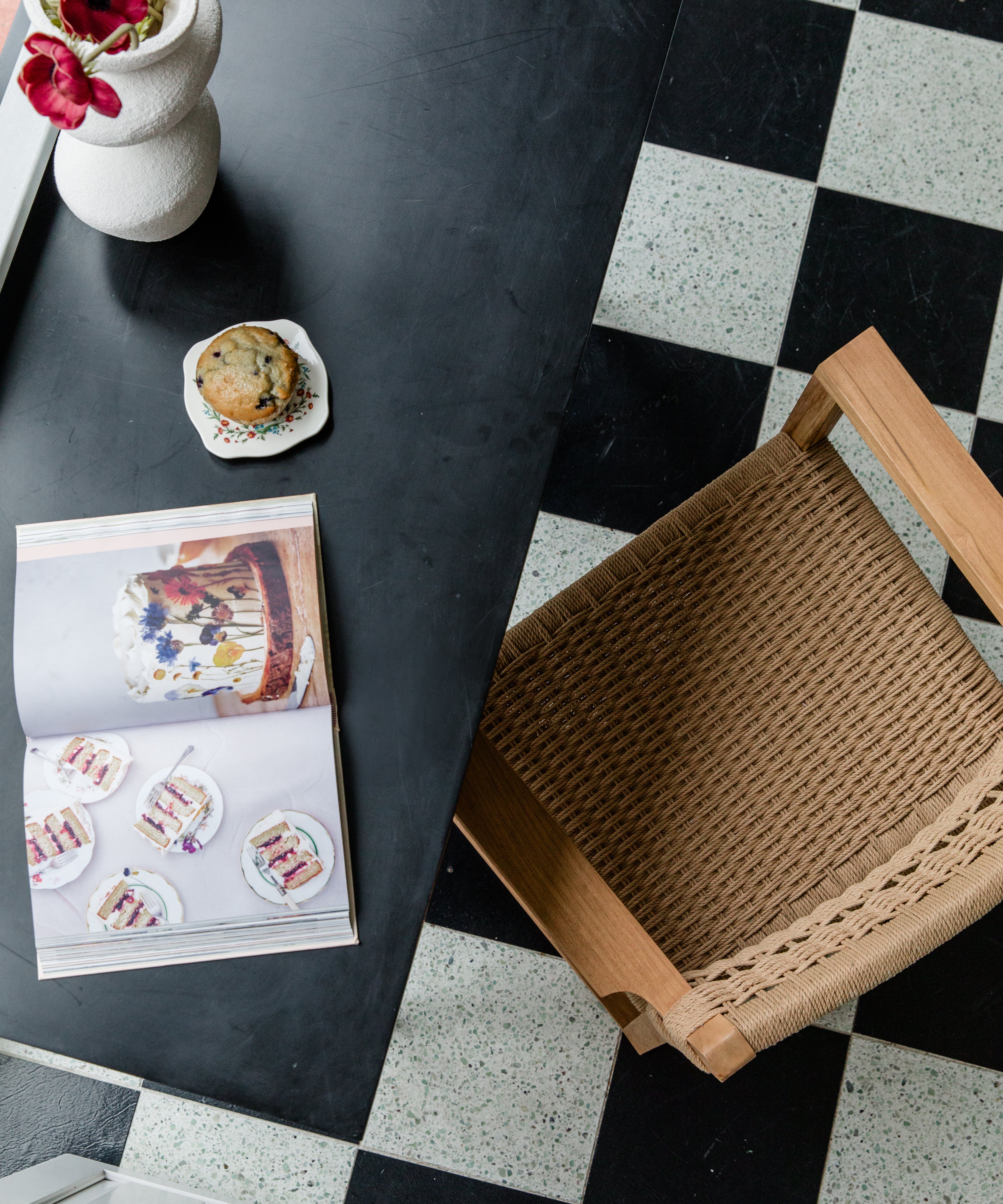 View of a cozy kitchen nook with a woven natural chair, flowers, a muffin, and cookbook