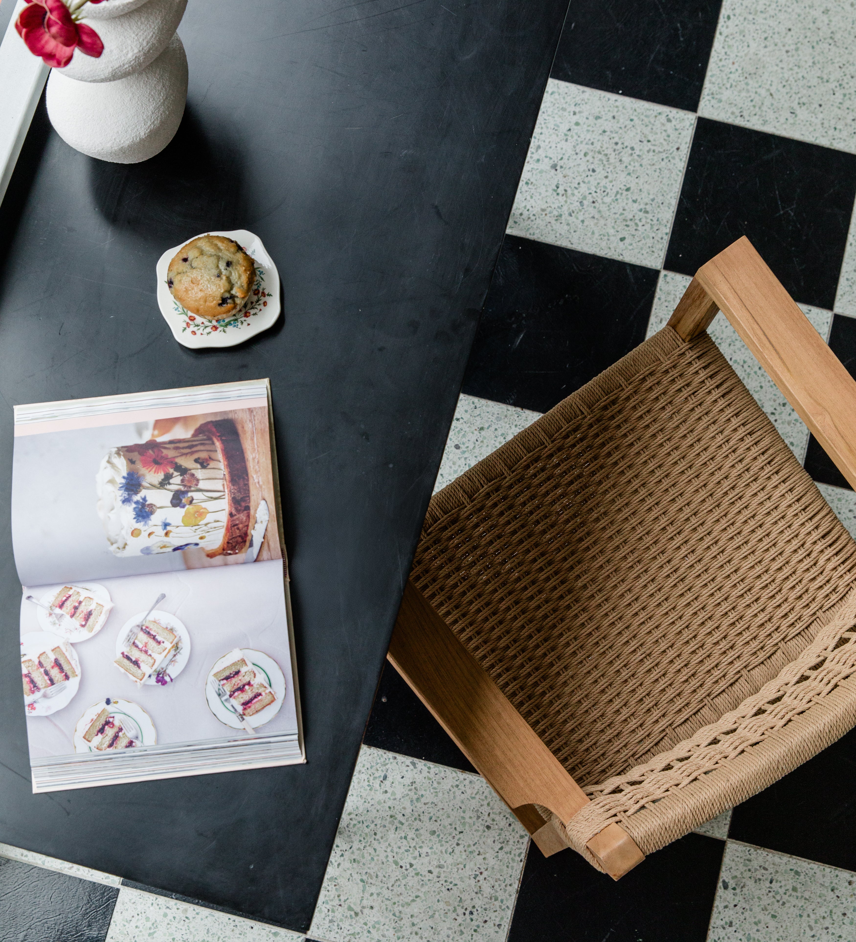 View of a cozy kitchen nook with a woven natural chair, flowers, a muffin, and cookbook